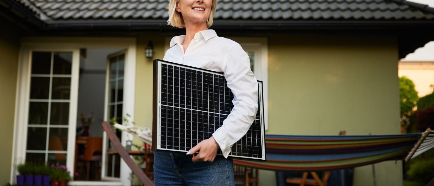 Lächelnde Frau hält ein Solarpanel vor einem Einfamilienhaus, Photovoltaik für Privathaushalte – TRIOSUN Energiesysteme
